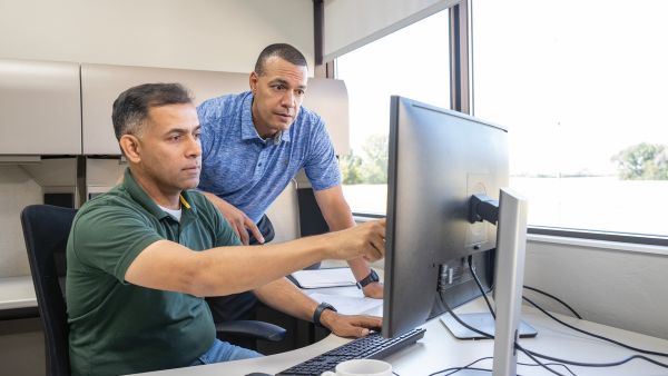 Two people in an office environment are looking at a large computer monitor. One person is seated and pointing at the screen, while the other stands beside the desk. The workspace includes cubicle partitions, a window with outdoor light, and a coffee cup on the desk.