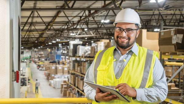 A person wearing a white safety helmet and a bright yellow reflective vest stands on an elevated platform inside a large warehouse. The individual is holding a tablet. The background shows rows of shelves stacked with boxes and packages under a high metal roof with industrial lighting.