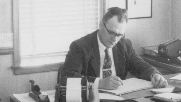 A black-and-white photo of Jack Keller seated at a desk in an office setting. The person is writing on a large sheet of paper. The desk holds a mechanical adding machine and other office supplies. A window with blinds and framed artwork are visible in the background.