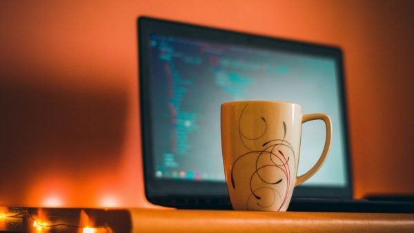 A ceramic coffee mug with abstract swirl designs sits on a desk in front of an open laptop displaying blurred code on the screen. Warm orange lighting creates a cozy atmosphere, and a string of small lights is partially visible on the left edge.